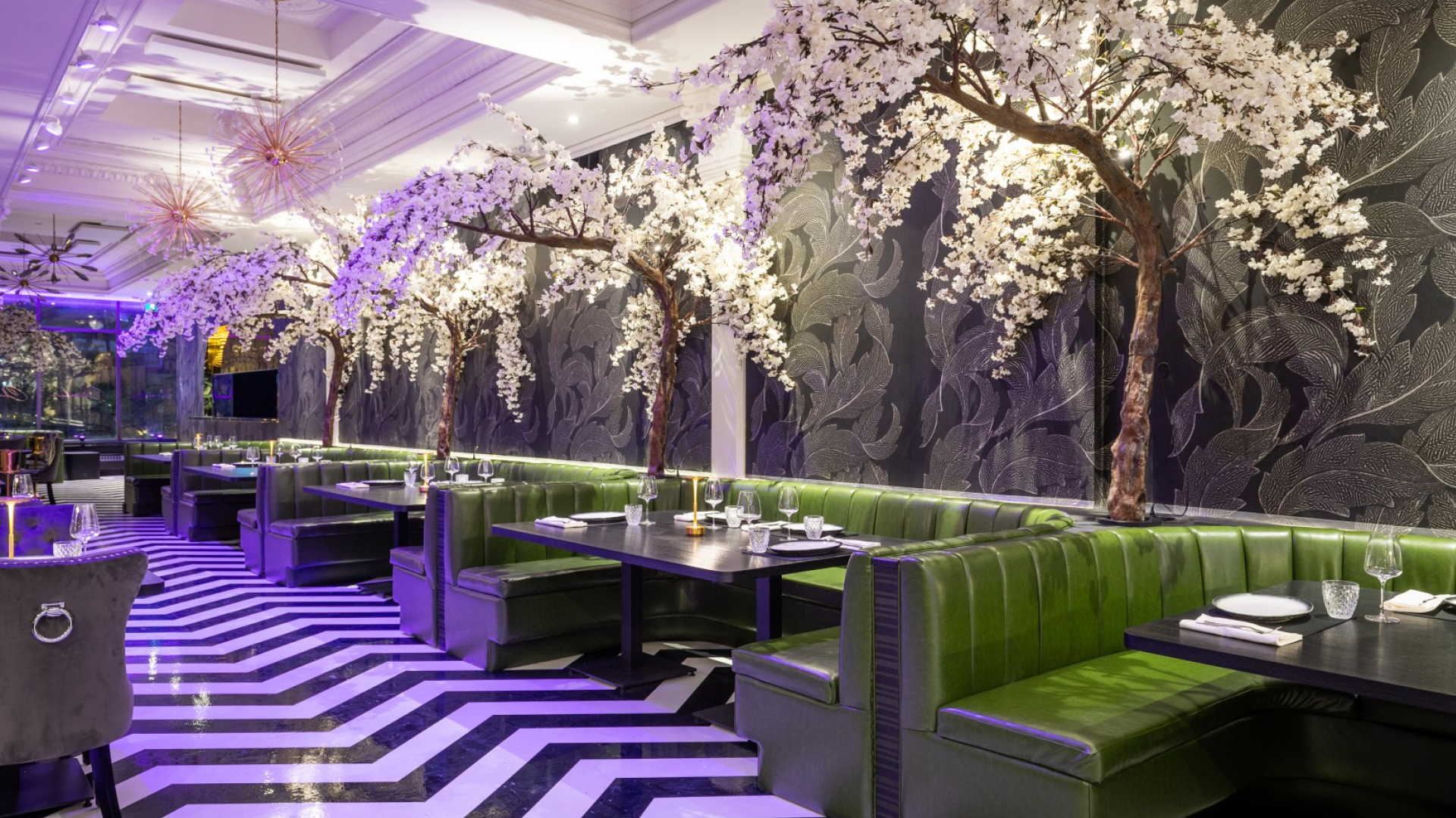 A restaurant dining area with green upholstered booths lining a zigzag black‑and‑white floor. Decorative flowering tree installations, glass chandeliers, and soft purple lighting create a dramatic, modern interior.